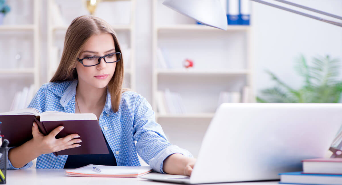 a woman in glasses holding a book in front of a laptop