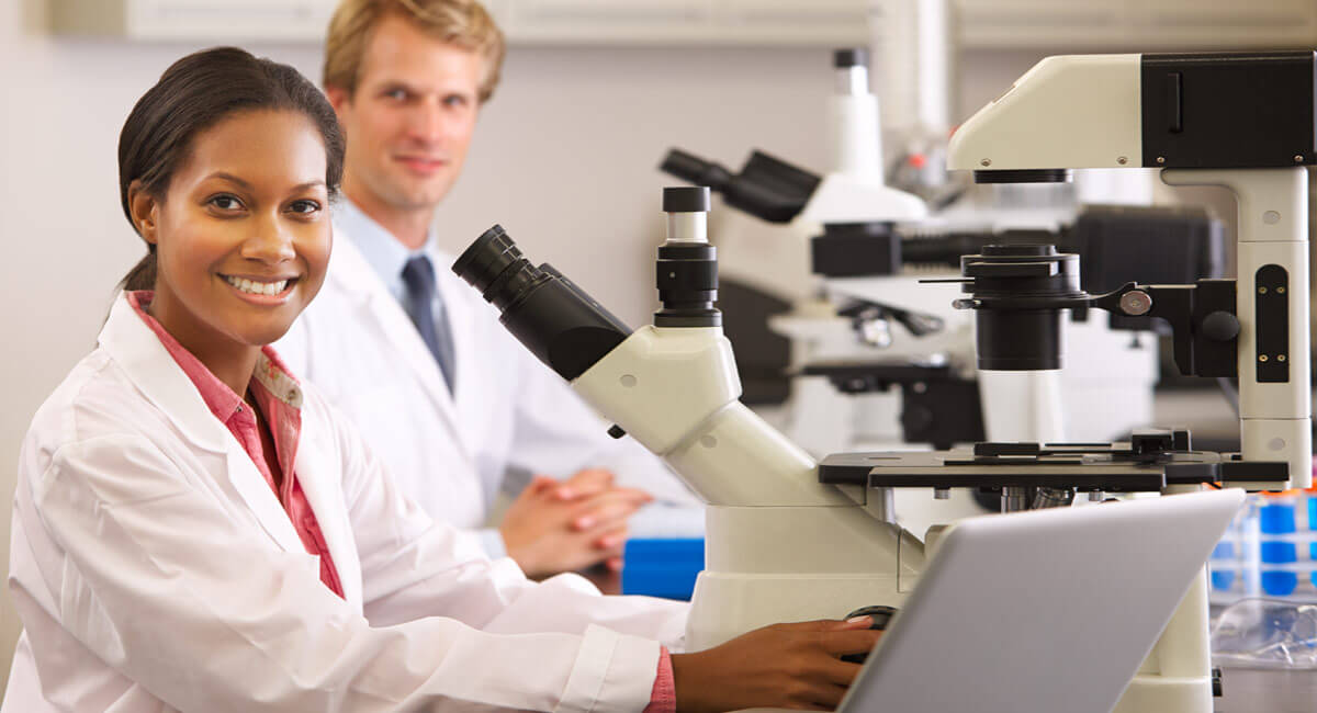 a woman smiling next to the microscope dressed in doctor's gown